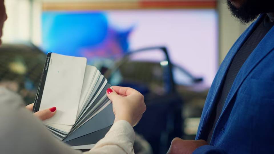 A woman holds a color palette while discussing options with a man in a car dealership, blurred cars visible in the background.