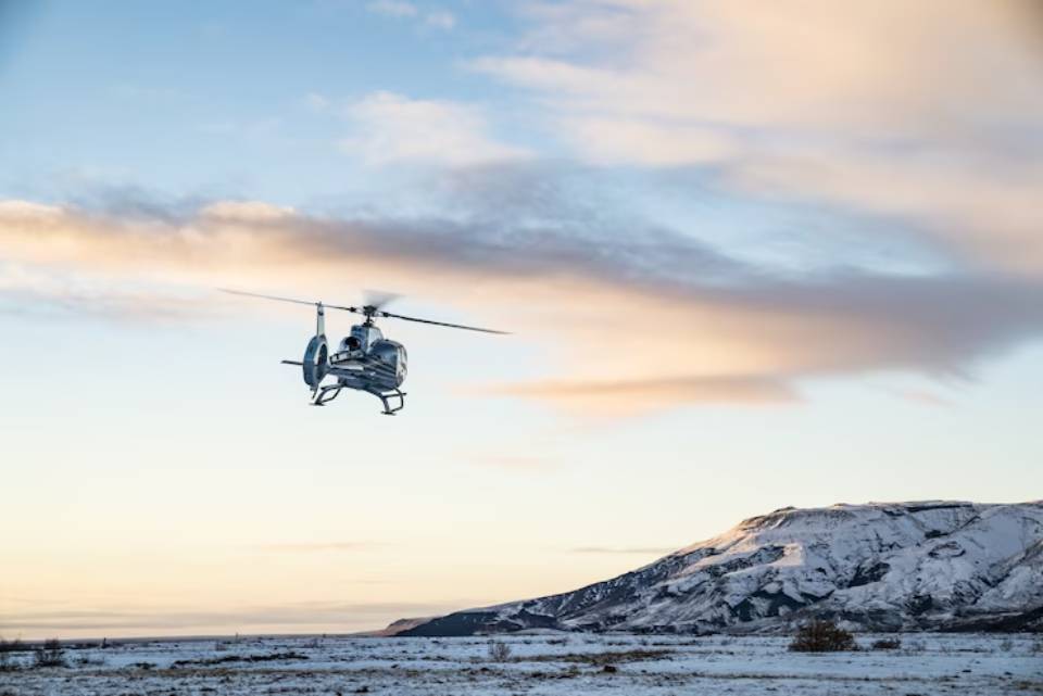 A helicopter flies over a snowy landscape at sunset, with colorful clouds and a distant mountain in the background.