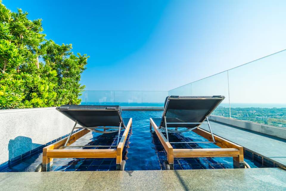 Two wooden lounge chairs beside a serene infinity pool, surrounded by greenery and overlooking a clear blue sky and distant landscape.