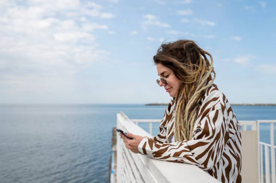 A person with long, dreadlocked hair is leaning on a white railing, focused on their phone against a serene ocean backdrop.