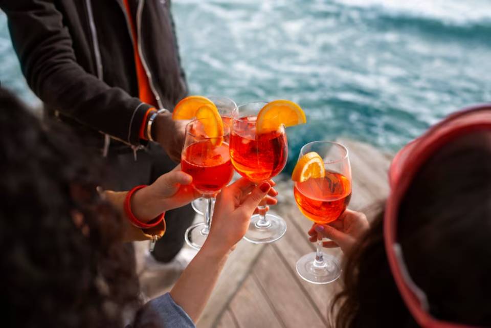A group of friends toast with colorful cocktails on a wooden dock, with a serene blue water backdrop and bright orange garnishes.