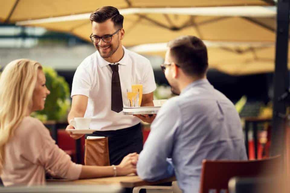 A waiter serves drinks at an outdoor café to two seated customers, one reaching for a cup, under a shaded awning.