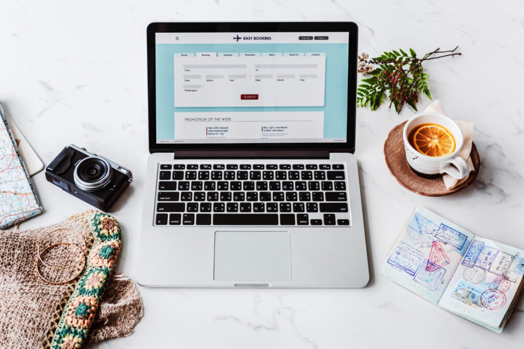 A laptop displaying a flight booking site, surrounded by a camera, travel map, passport with stamps, and a cup of tea with orange slice.