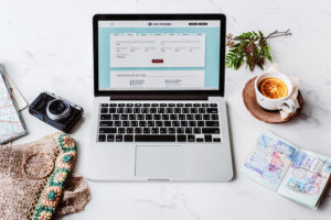 A laptop displaying a flight booking site, surrounded by a camera, travel map, passport with stamps, and a cup of tea with orange slice.