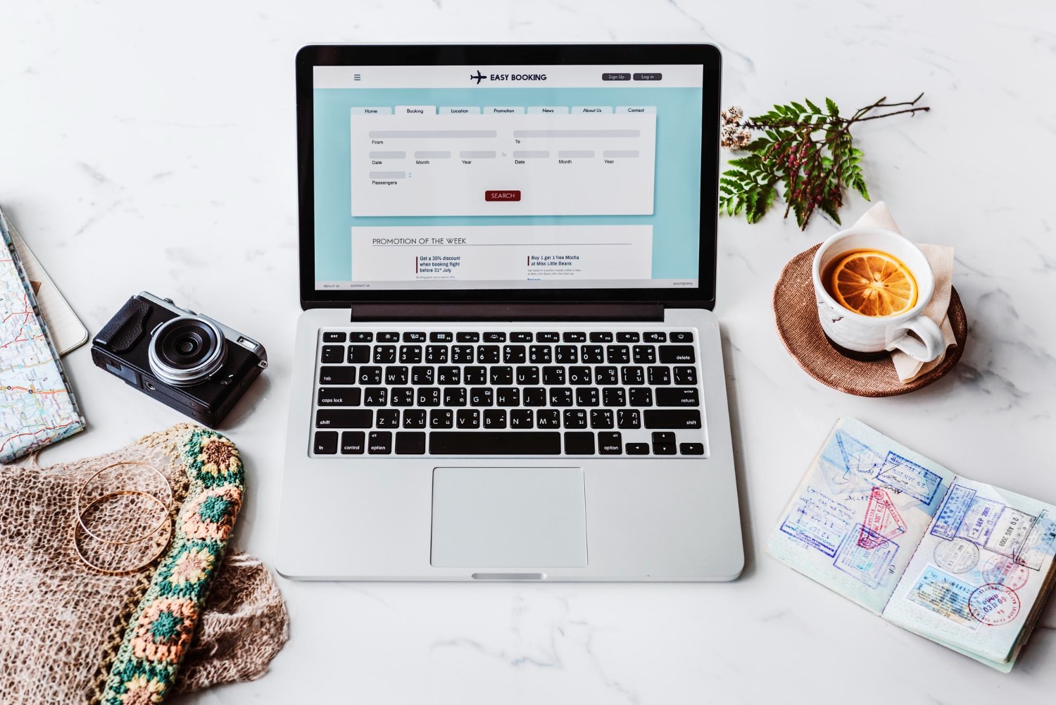 A laptop displaying a flight booking site, surrounded by a camera, travel map, passport with stamps, and a cup of tea with orange slice.