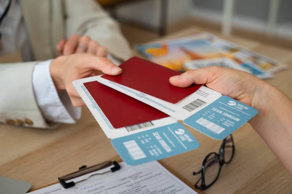 A hand passes red passports and blue airline tickets across a wooden desk, with travel brochures and glasses in the background.