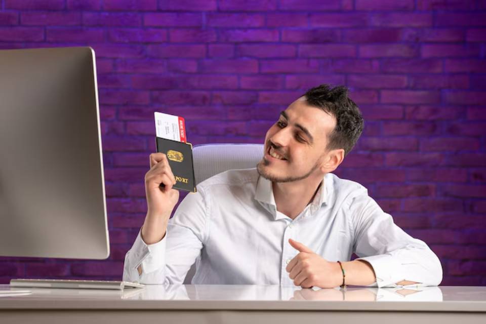 A person in a white shirt holds a passport and credit card while sitting at a desk in front of a computer, with a purple brick background.