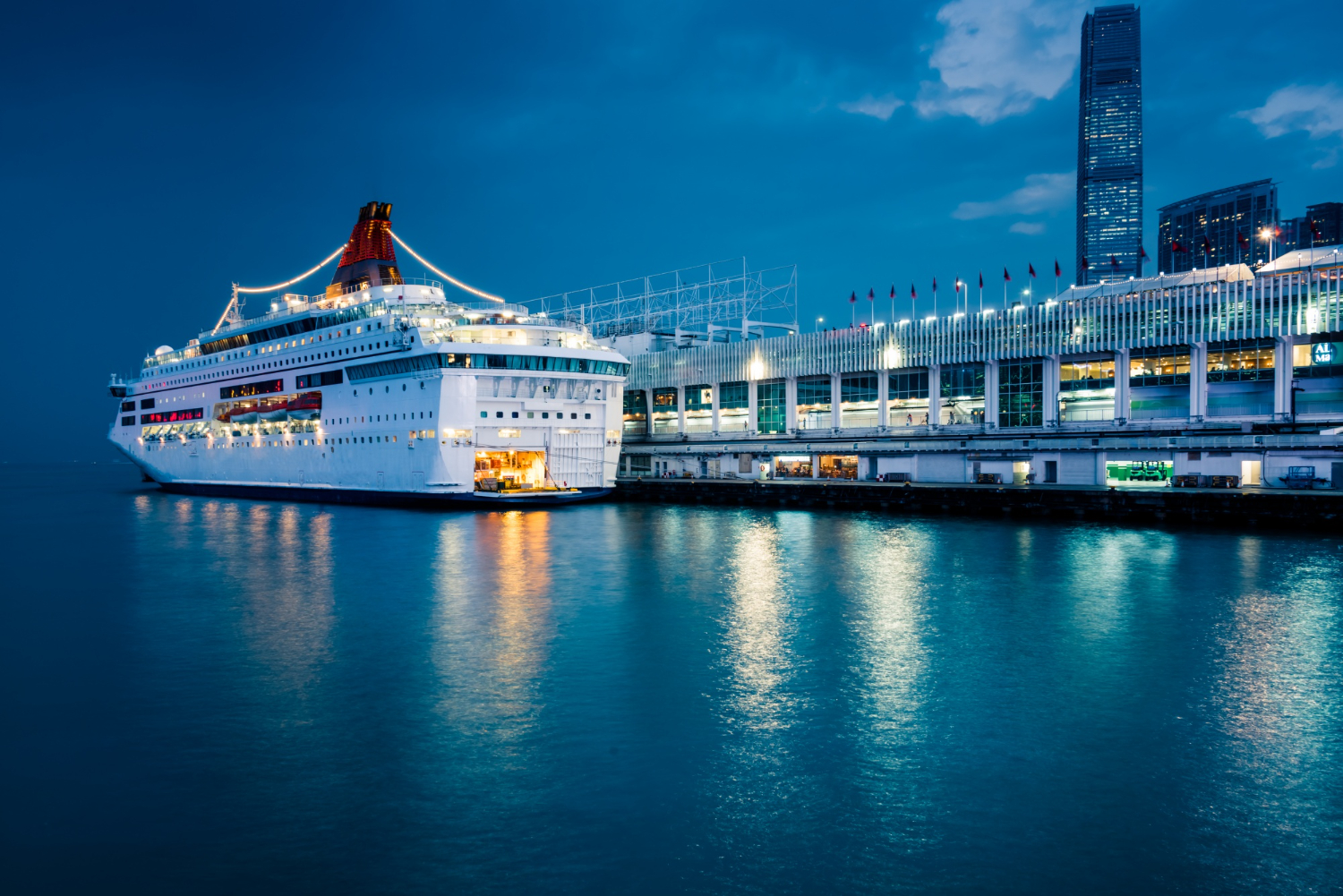 A large cruise ship docked at night beside a bustling terminal, with city lights reflecting on the calm water.