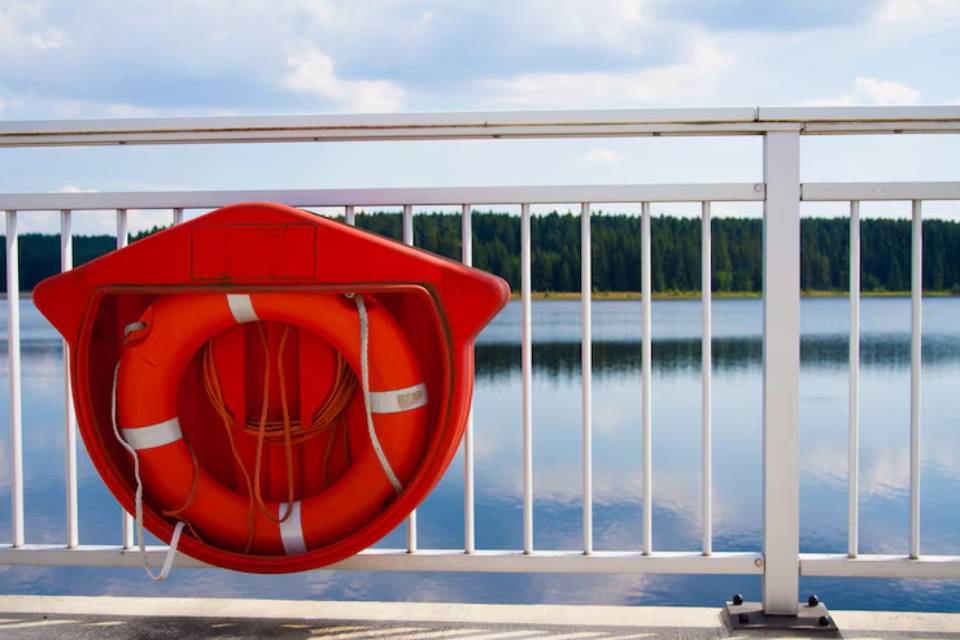 A bright orange life ring secured to a white railing, with a calm lake and lush trees reflected in the water behind it.