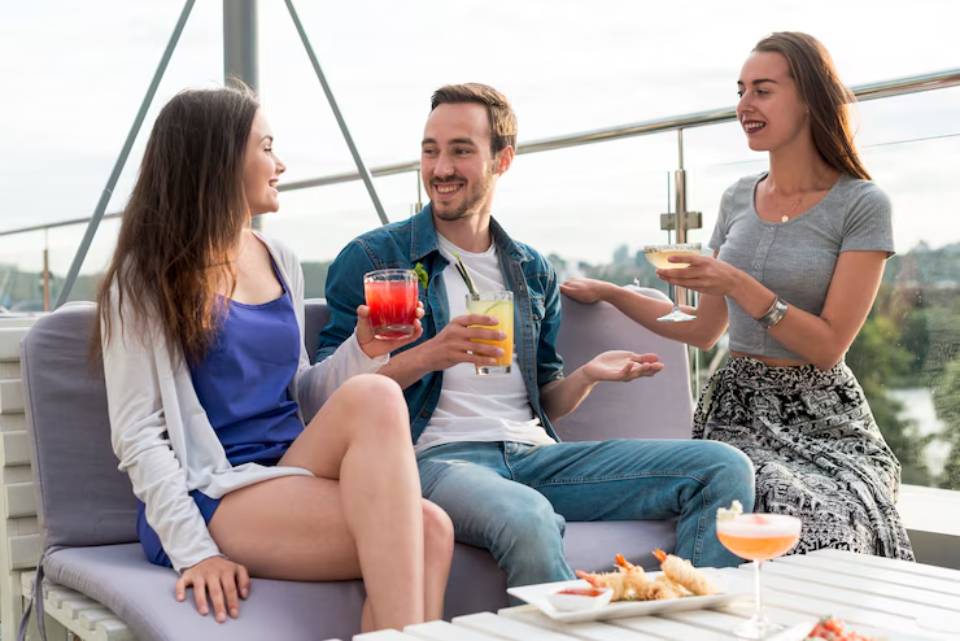Three friends enjoy vibrant cocktails on a modern rooftop lounge, with a scenic view in the background and plate of appetizers on the table.