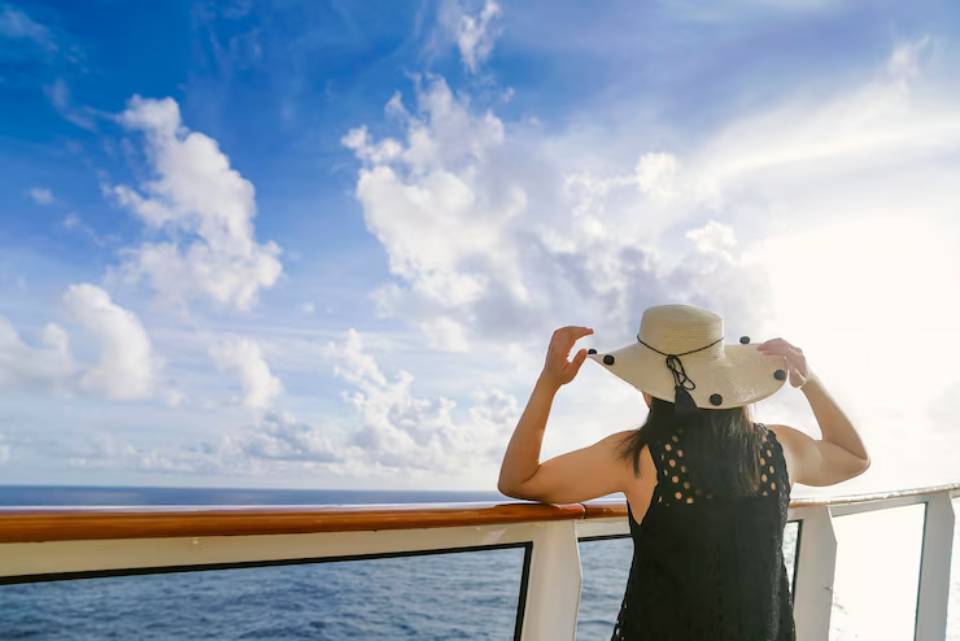 A woman in a black dress and wide-brimmed hat stands on a cruise ship, gazing at the blue ocean and dramatic sky filled with clouds.