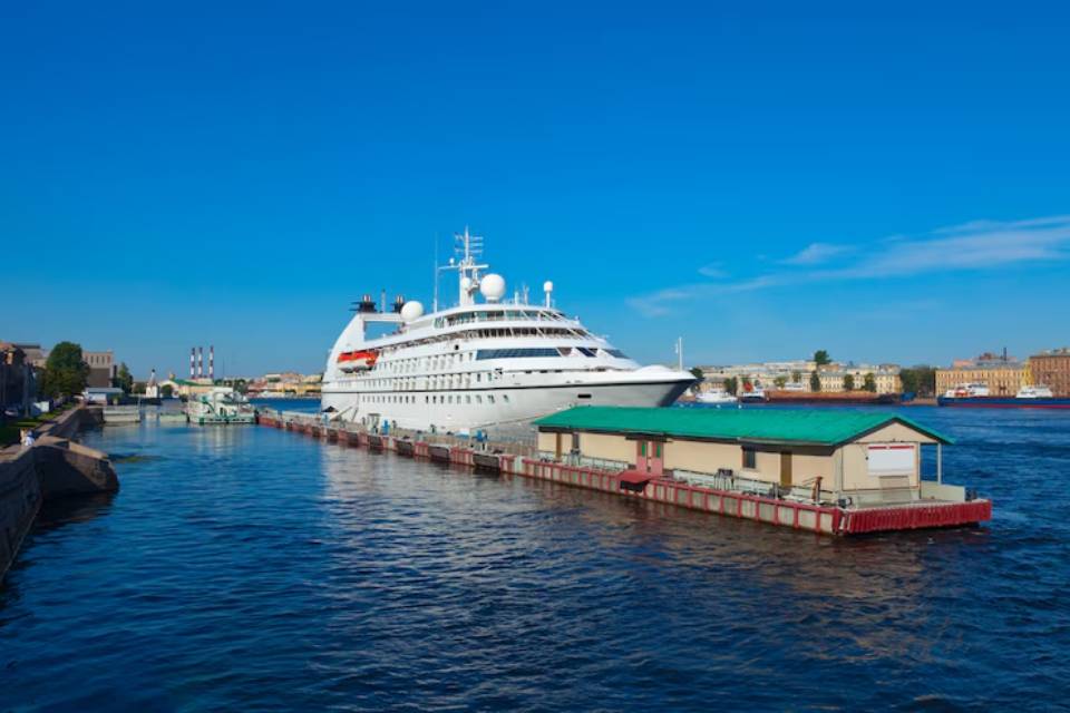 A large white cruise ship docked at a riverside pier under a clear blue sky, surrounded by historic buildings.