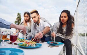 Close friends enjoy sushi on a sunny day, surrounded by drinks and snacks on a white table by a fence.