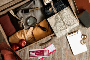 An open suitcase filled with clothes, a magnifying glass, a map, apples, boarding passes, and a clipboard on a textured surface.