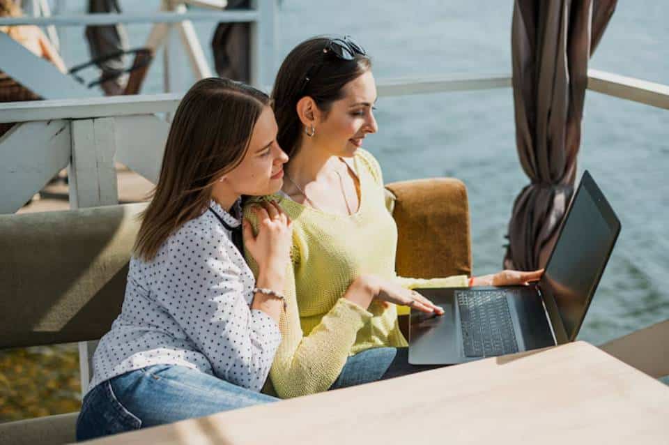 Two women sit on a cozy sofa by the water, one working on a laptop while the other leans in closely, sharing the moment.
