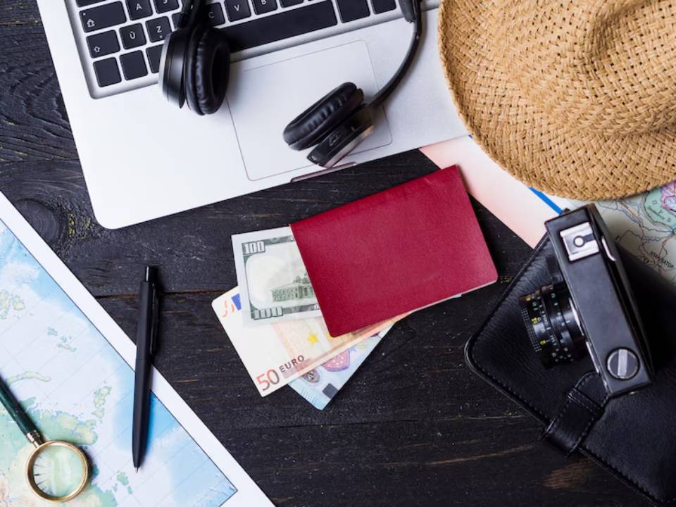 Travel essentials arranged on a wooden desk: laptop, headphones, camera, passport, cash, maps, and a sunhat.