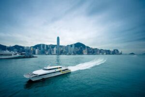 A sleek ferry navigates through Hong Kong's harbor, with towering skyscrapers and a mountainous backdrop under a cloudy sky.