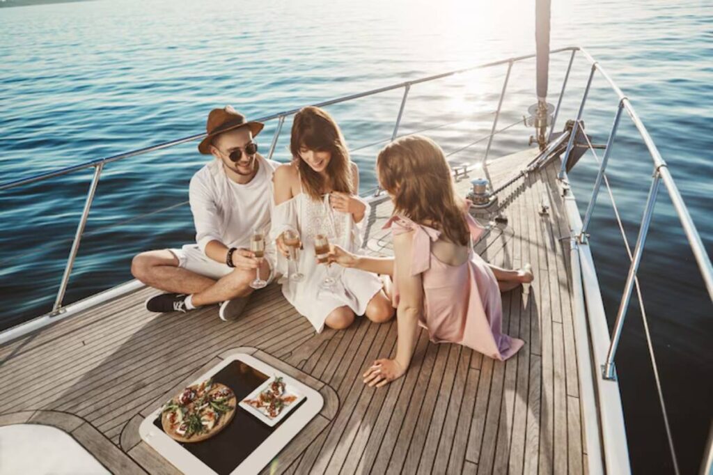 Three friends on a yacht toast with drinks, enjoying a sunny day over a spread of food on a wooden deck beside calm waters.