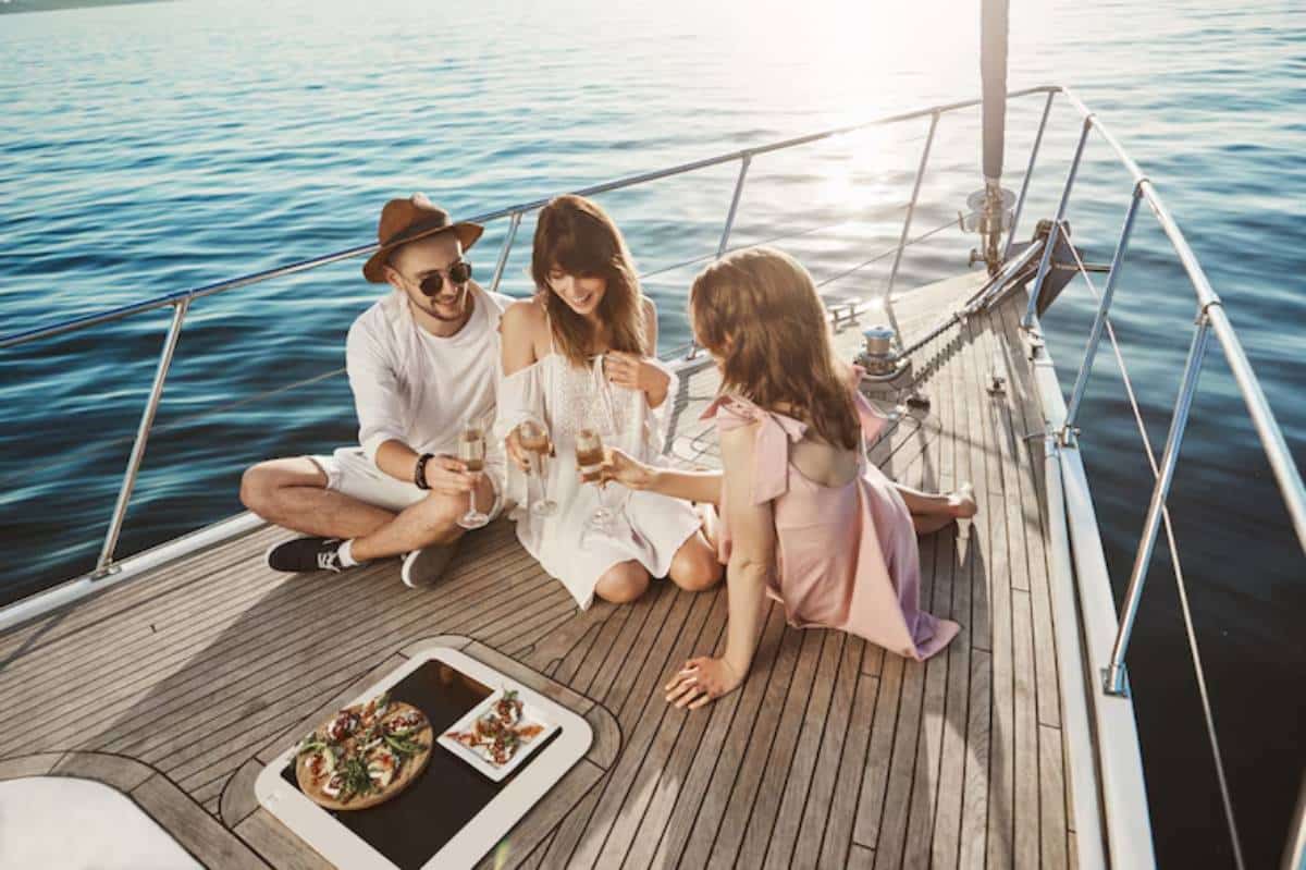 Three friends on a yacht toast with drinks, enjoying a sunny day over a spread of food on a wooden deck beside calm waters.