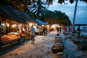 A bustling beach market at dusk, lined with illuminated stalls selling fruits and goods, with palm trees and a tranquil sea in the background.