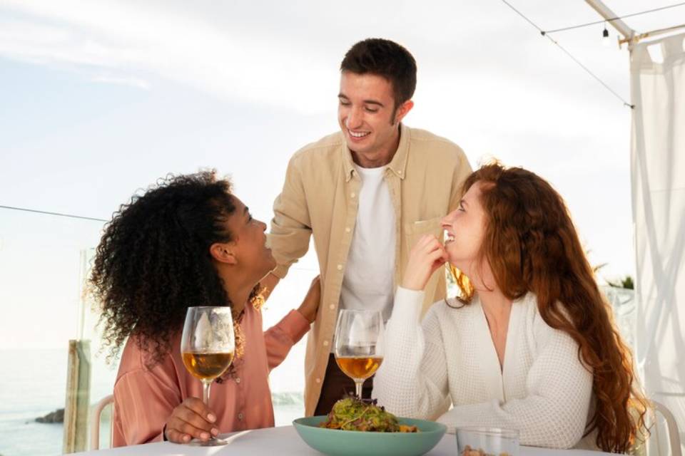 A casual outdoor lunch setting with three friends enjoying drinks and a beautiful view of the ocean in a relaxed atmosphere.
