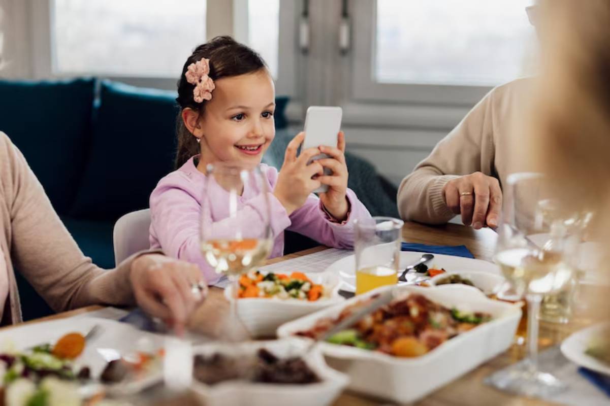 A festive family meal featuring a variety of dishes on a wooden table, with drinks and a child using a smartphone.
