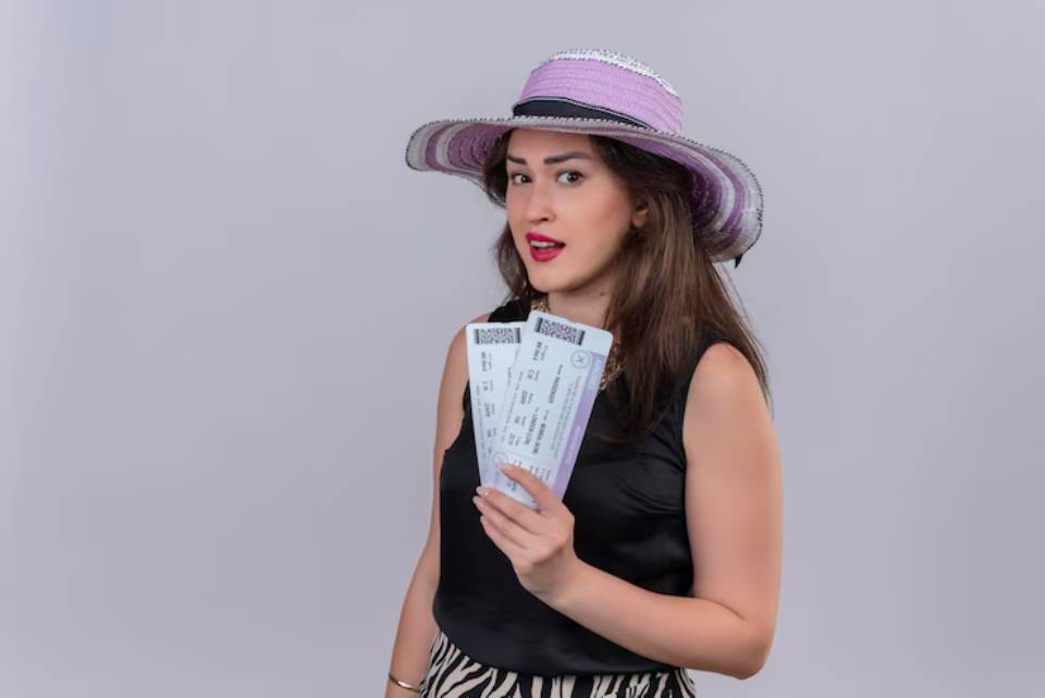 A woman wearing a large purple hat holds three flight tickets, showcasing excitement for her upcoming travel plans.