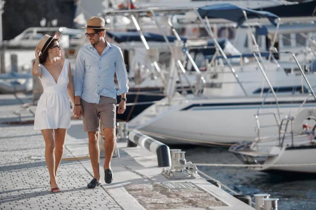 A couple walks hand in hand along a sunny marina, surrounded by yachts and boats, exuding a relaxed vacation vibe.