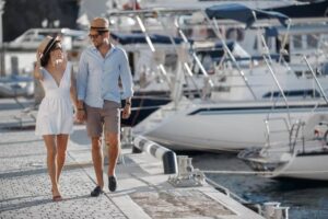 A couple walks hand in hand along a sunny marina, surrounded by yachts and boats, exuding a relaxed vacation vibe.