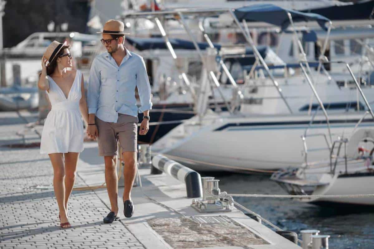 A couple walks hand in hand along a sunny marina, surrounded by yachts and boats, exuding a relaxed vacation vibe.