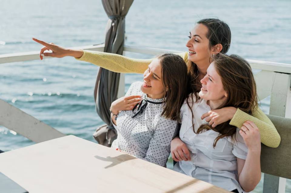 Three women sit together by the water, with one pointing excitedly into the distance while enjoying the scenic backdrop.