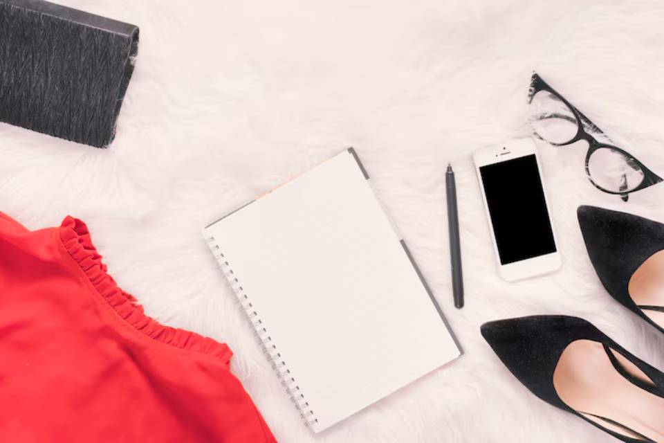 Flat lay of a red ruffled top, black heels, a notebook, smartphone, pen, glasses, and a black clutch on a soft white background.