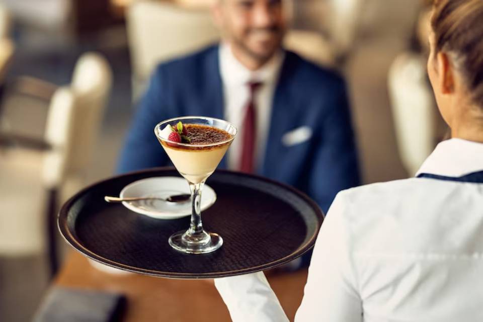 A waiter presents a dessert glass filled with custard, topped with caramel and garnished with berries, on a round tray in a restaurant setting.