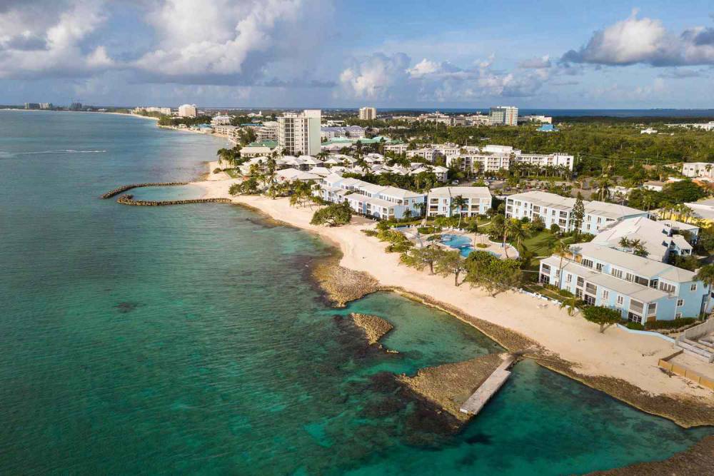 Aerial view of a tropical coastline with sandy beaches, clear turquoise waters, palm trees, and resort buildings under a partly cloudy sky.