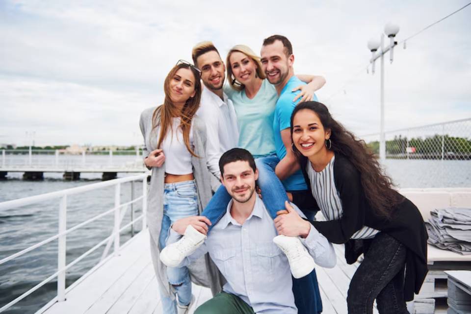 A group of seven friends poses playfully on a boat dock, enjoying a fun day by the water with a cloudy sky overhead.