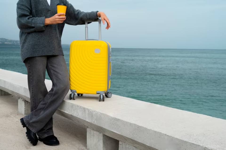 A person in a gray sweater holds a yellow cup beside a bright yellow suitcase on a coastal walkway, with the ocean in the background.
