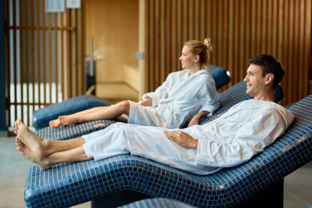 Two individuals in white bathrobes relax on stylish blue lounge chairs in a tranquil spa setting, surrounded by wooden accents.