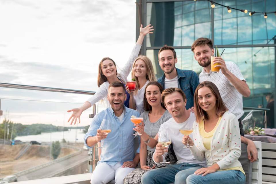 A group of friends joyfully posing with colorful cocktails on a balcony with a scenic view and festive atmosphere.