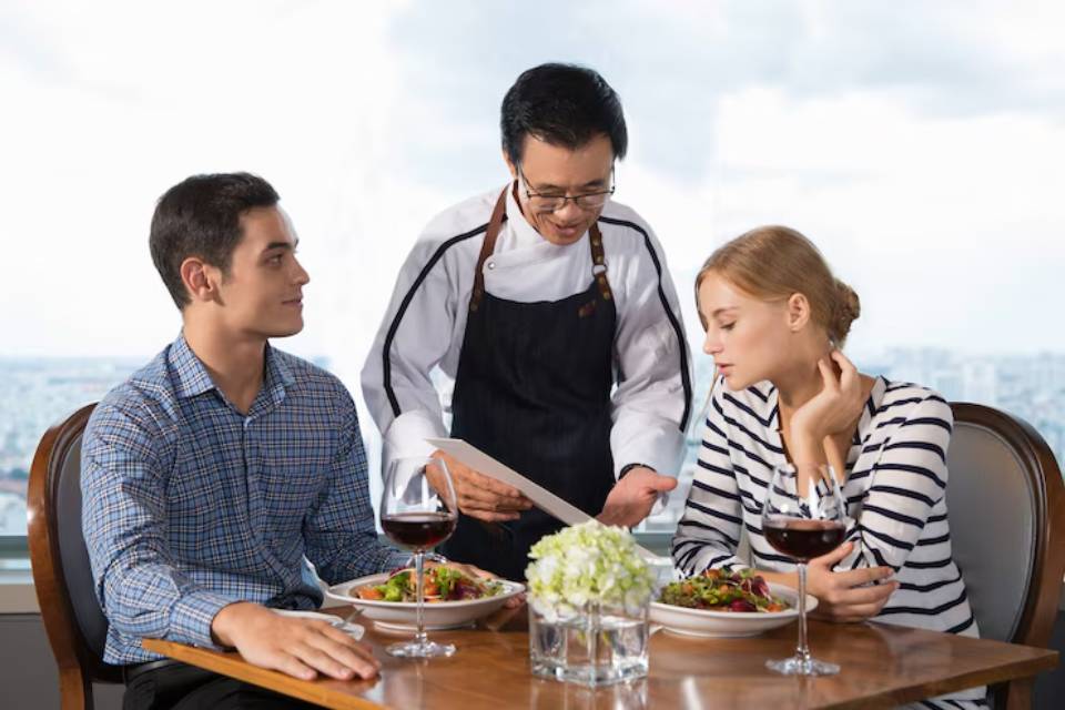 A waiter in an apron presents a menu to a couple dining at a table with gourmet dishes and red wine, with a city skyline view.