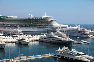 A bustling marina featuring a large cruise ship alongside various luxury yachts docked in bright blue water under a clear sky.
