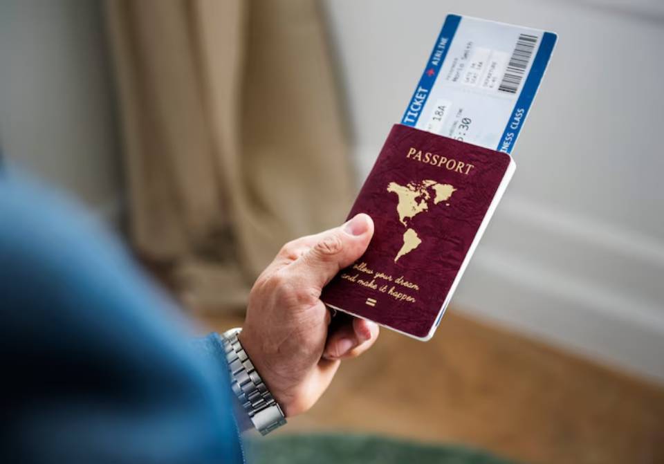 A person's hand holding a maroon passport with a world map design, accompanied by a blue airline ticket in a travel setting.