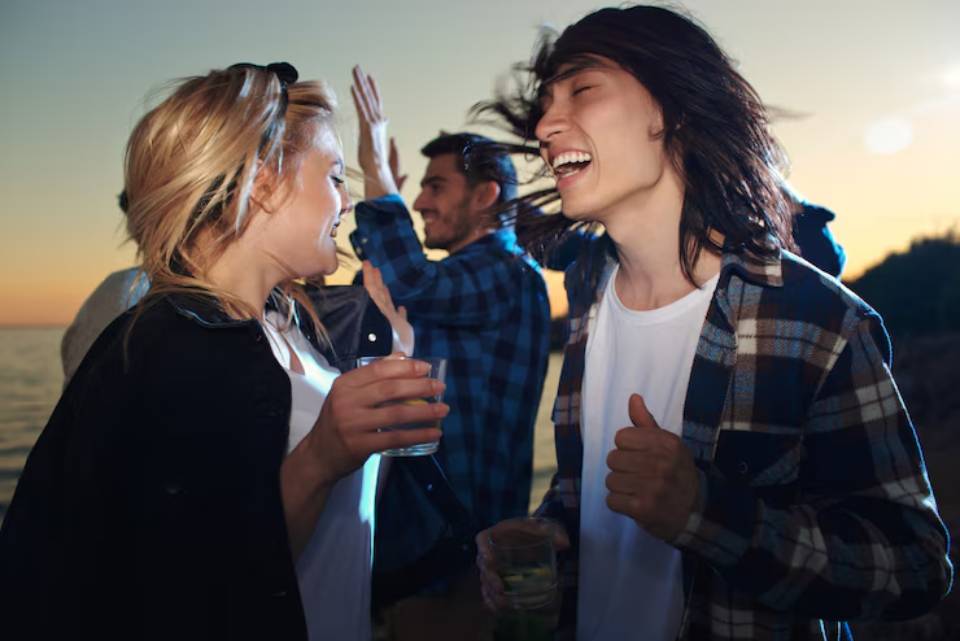 A group of young people enjoying a sunset beach party, laughing and holding drinks while celebrating together.