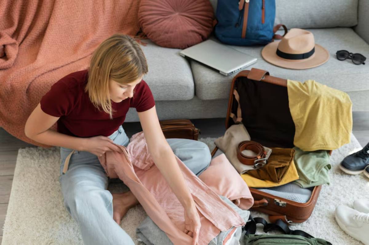 A person sorts through clothes while packing a suitcase, surrounded by bags, a laptop, sunglasses, and a hat. Cozy living room setting.