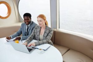 A professional setting with two people working on a laptop at a circular table, enjoying a drink with an ocean view visible through the window.