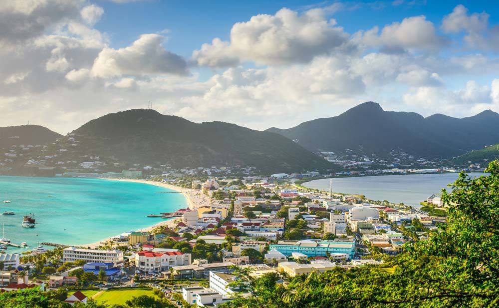 A panoramic view of St. Maarten featuring turquoise waters, a vibrant shoreline, colorful buildings, and surrounding green mountains.