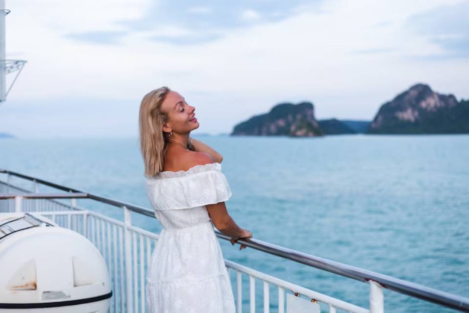 A woman in a white off-shoulder dress stands by the railing on a boat, gazing at lush islands and calm blue waters.