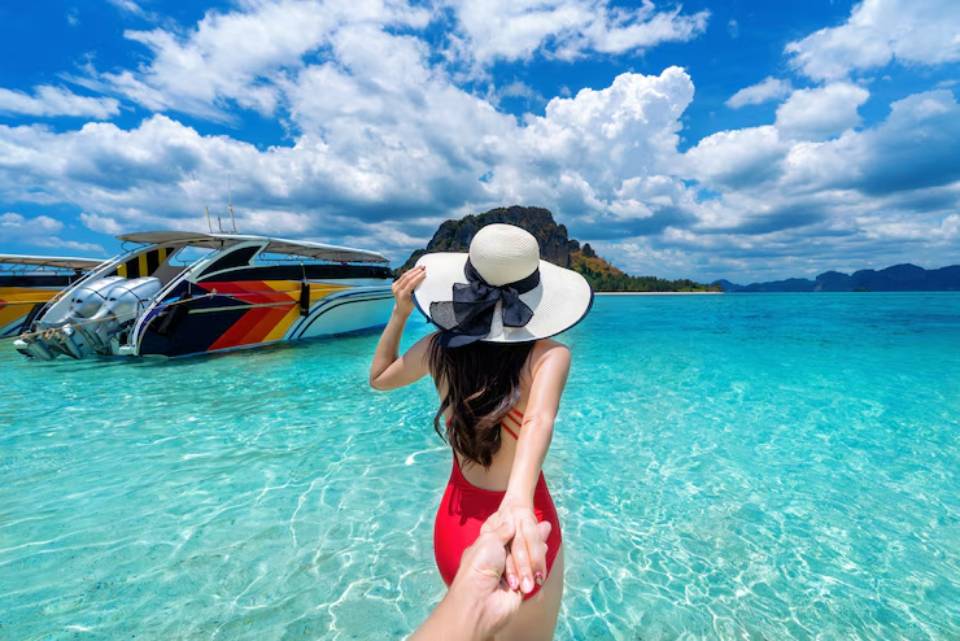A woman in a red swimsuit and large sun hat holds hands with someone, standing in crystal-clear water by colorful boats under a blue sky.