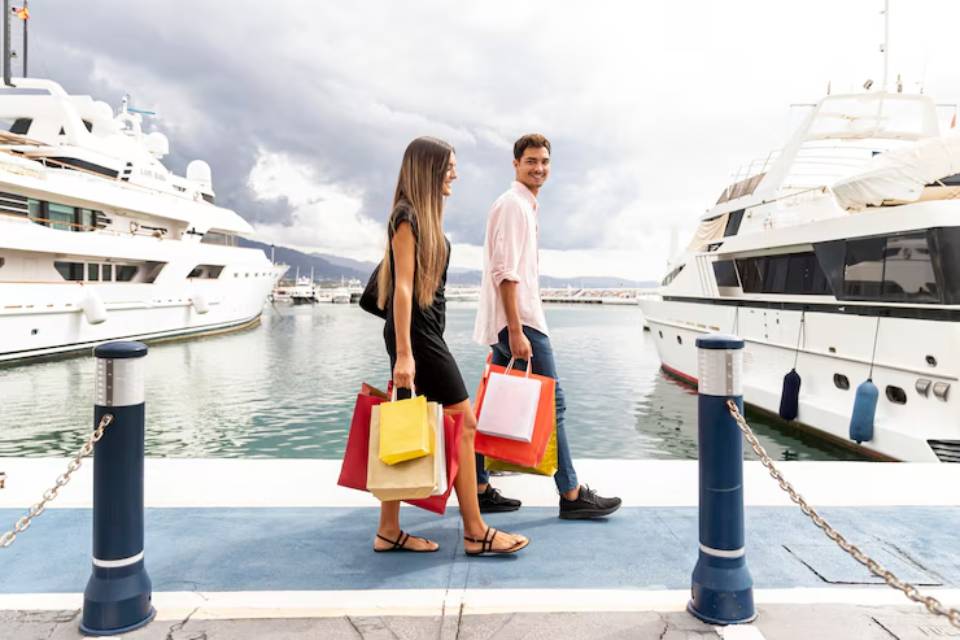 A couple strolls along a marina, each carrying colorful shopping bags beside luxury yachts under a cloudy sky.