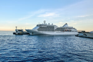 A large white cruise ship docked at a pier, surrounded by calm blue water and a clear sky.