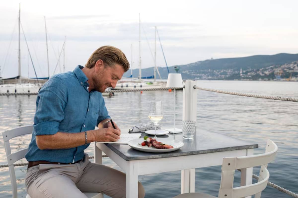 A man in a blue shirt sits at a table by the water, writing notes beside a plate of food and a glass of wine. Sailboats are in the background.